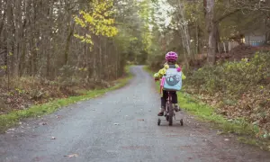 Dziecko w kasku jedzie na rowerze z bocznymi kółkami po leśnej ścieżce.