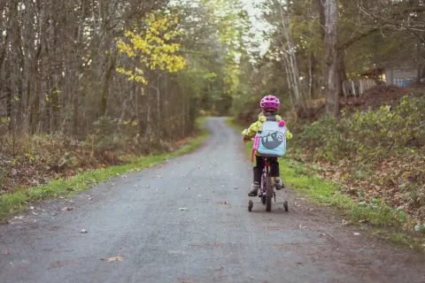 Dziecko w kasku jedzie na rowerze z bocznymi kółkami po leśnej ścieżce.